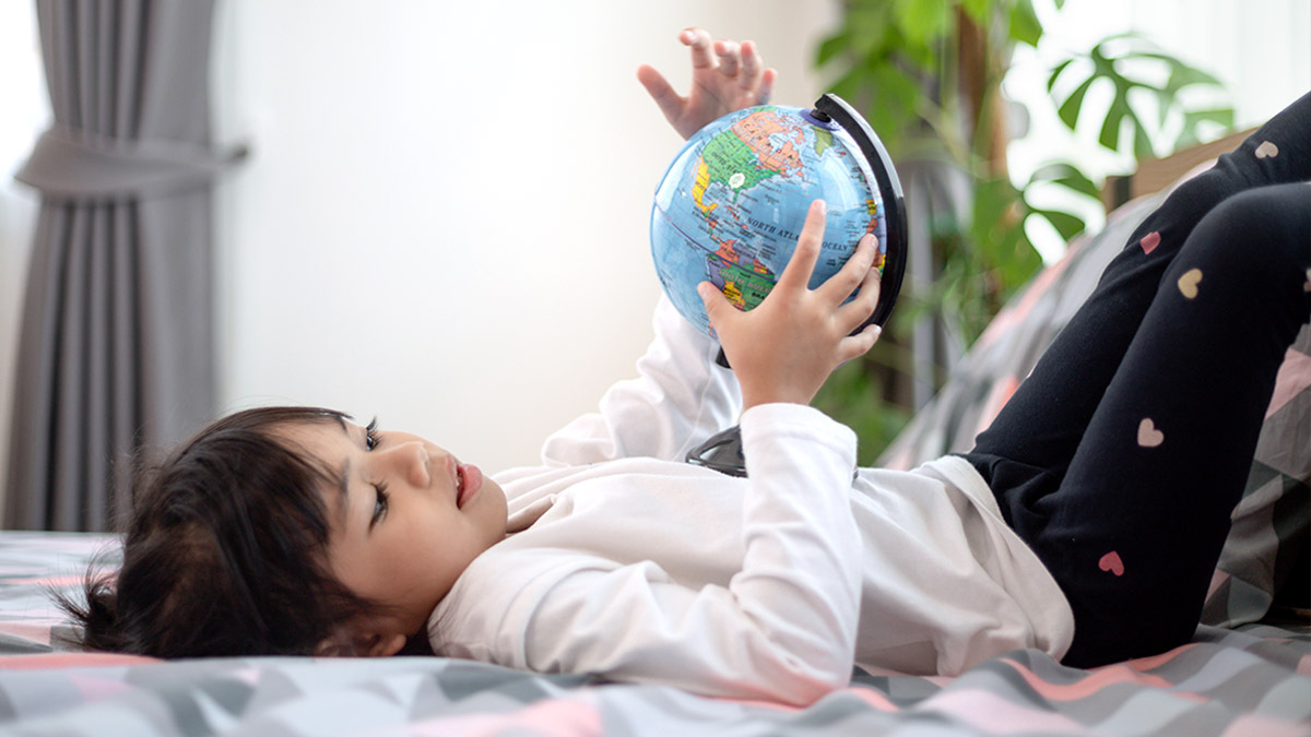 A young child lies on his bed and holds a small globe in their hands