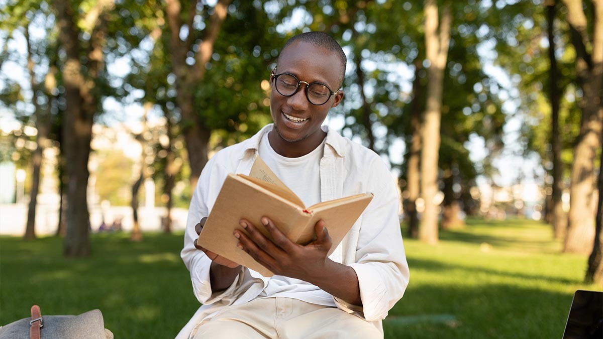 A young man with glasses reads a book in the park