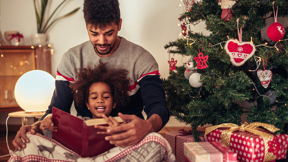 A father reads the Bible to his daughter by their Christmas tree