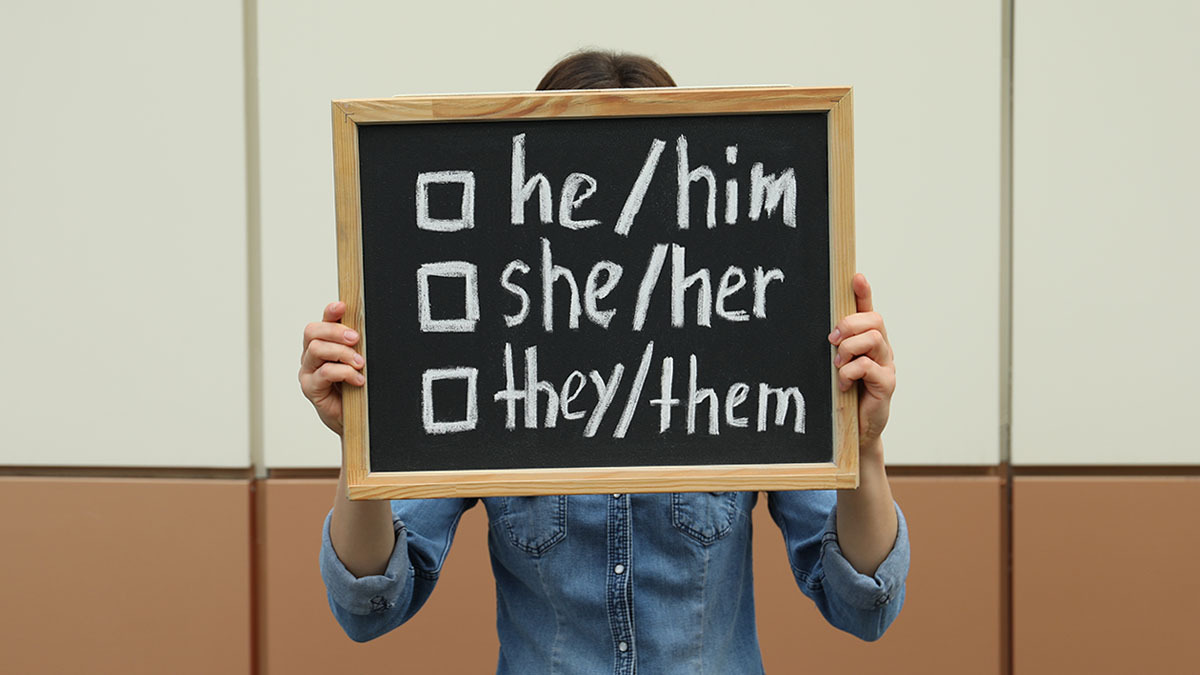 A young girl holds up a sign with a list of personal pronouns: he/him, she/her, and they/them