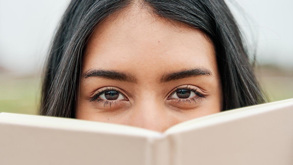 A teen girl peeks out from behind the cover of a book