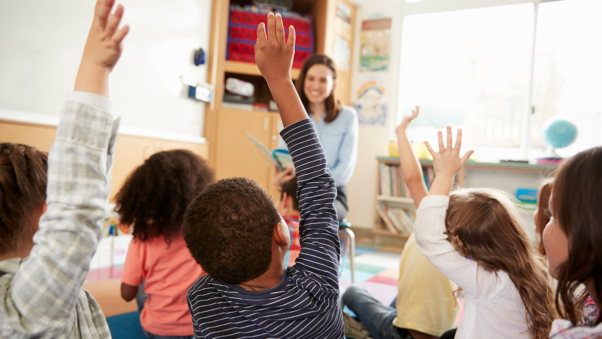 A class of children sit on a matt and raise their hands for the teacher