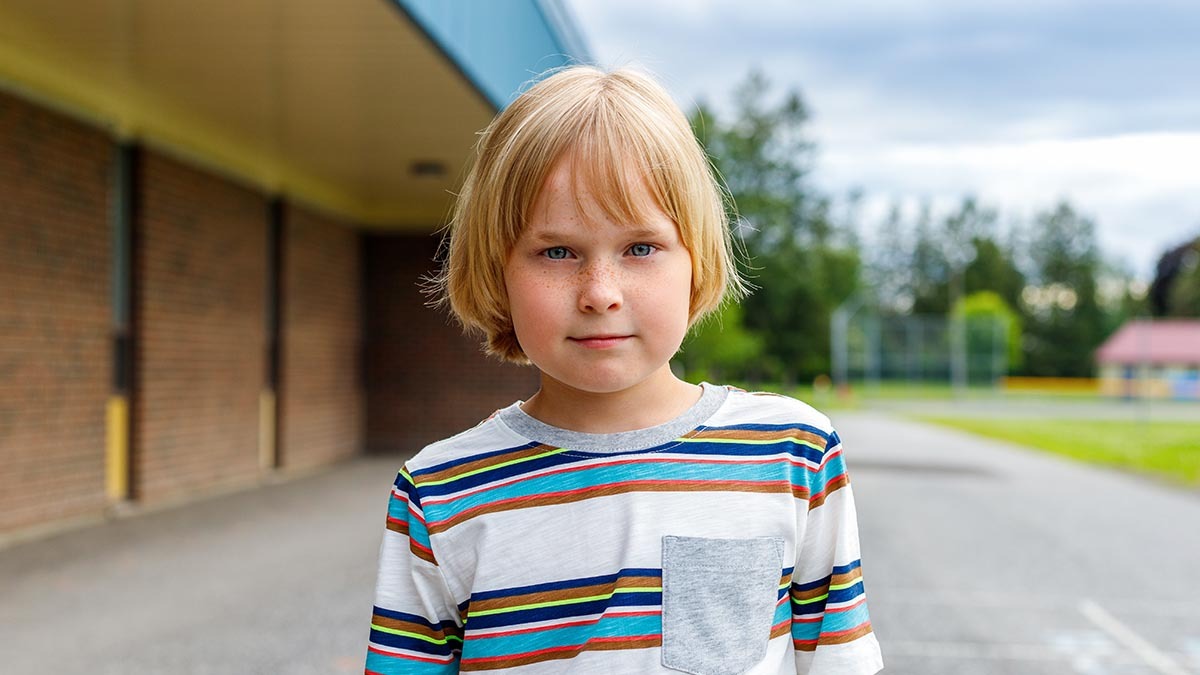 A young boy looks at the camera, a school yard is in the background