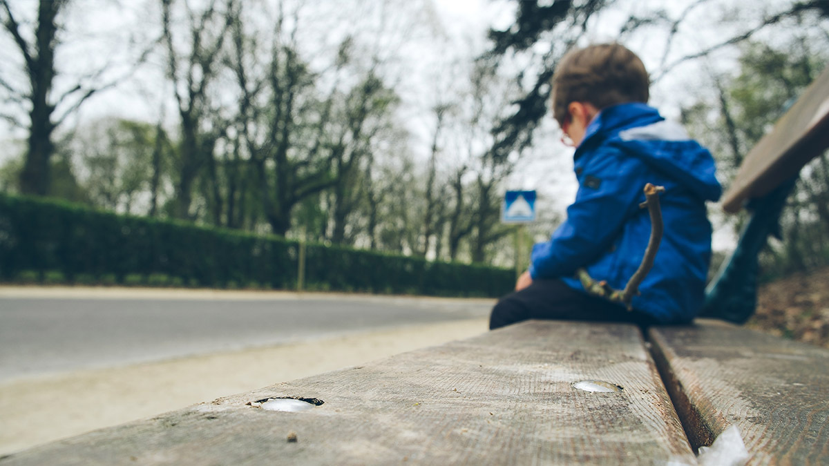 A boy sits on the end of a park bench and looks downcast