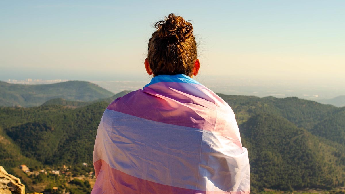 A person wearing a transgender pride flag around their shoulders looks out into the horizon