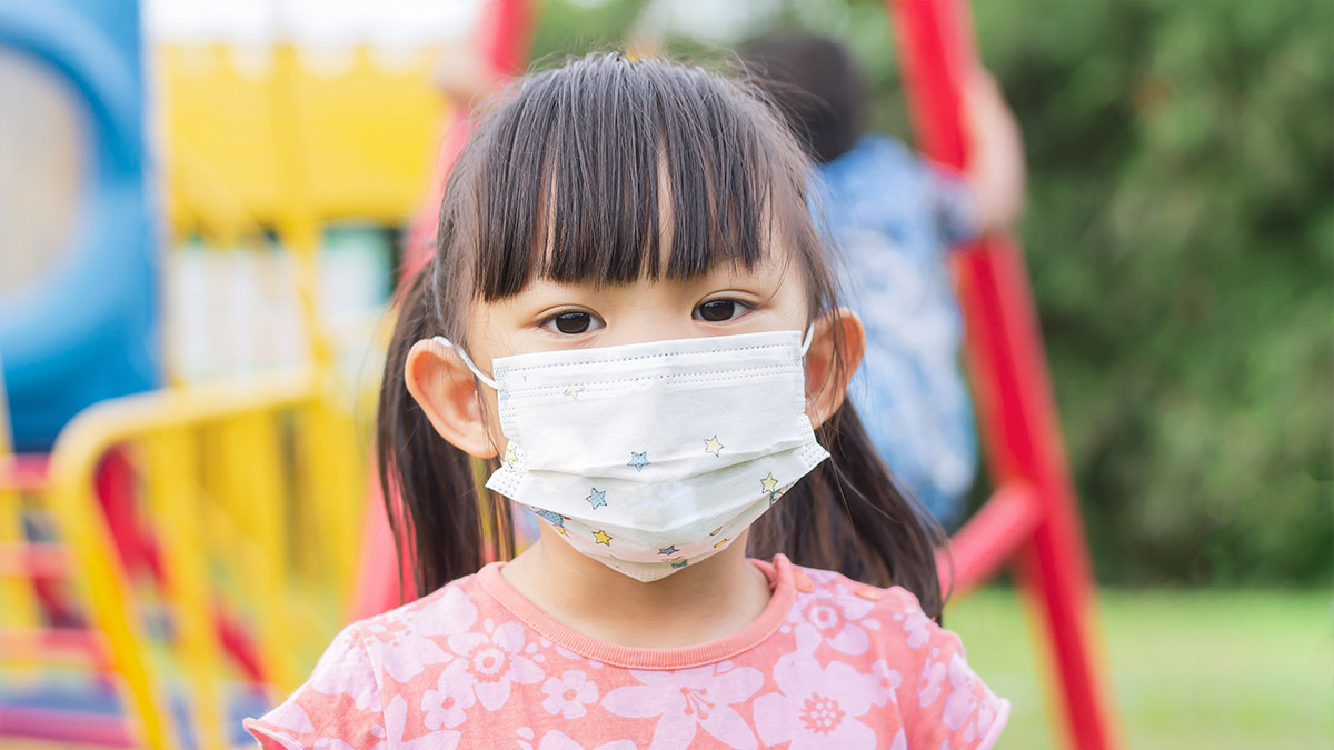 A young girl wearing a medical mask peeks over a fence