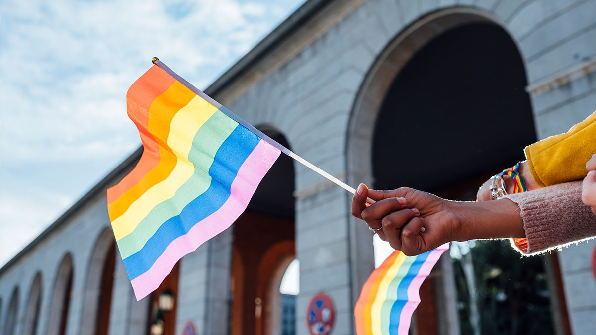 A hand holds up a gay pride flag outdoors in a city