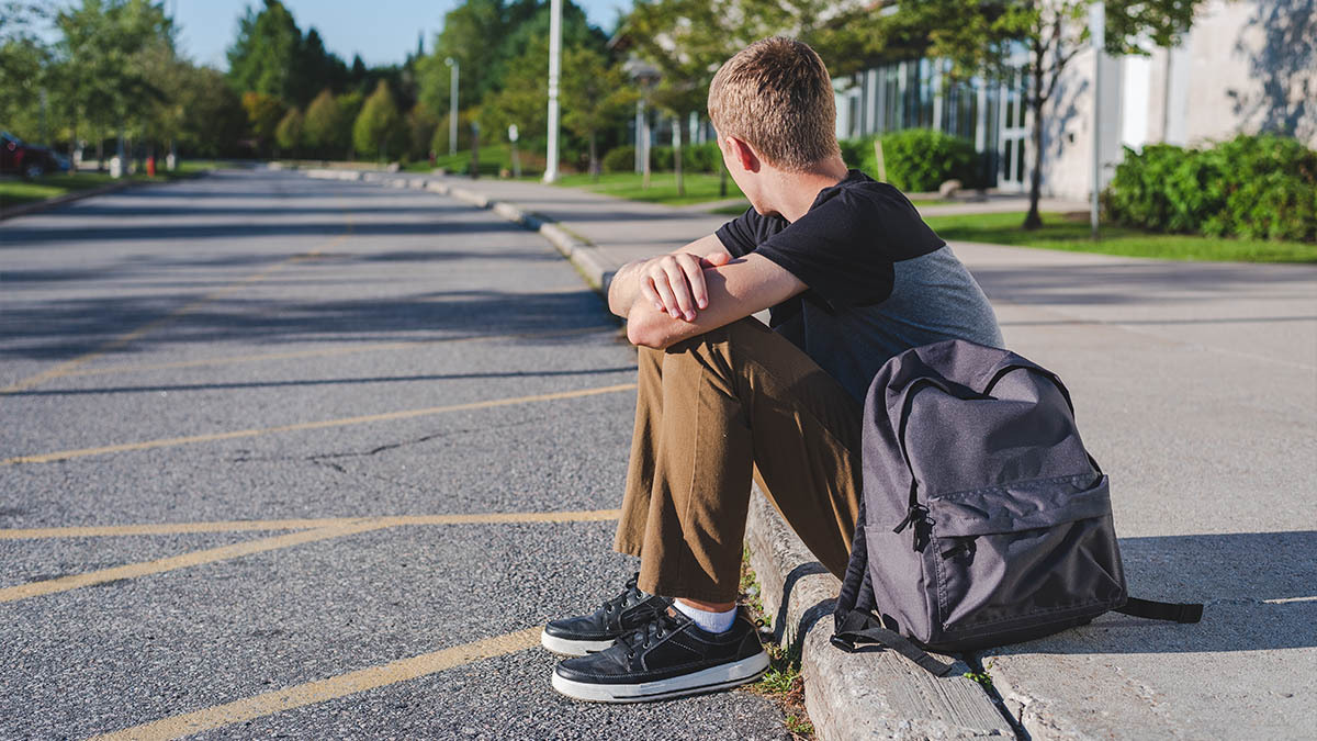 A teen boy sits on the curb with his backpack and looks away