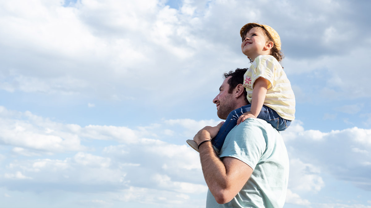 A father carries his little daughter on his shoulders as she looks at the sky