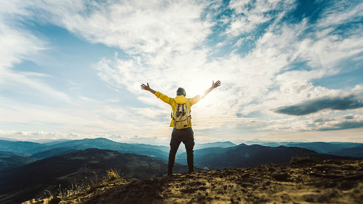 A man stands on top of a mountain and raises his hands towards the sun