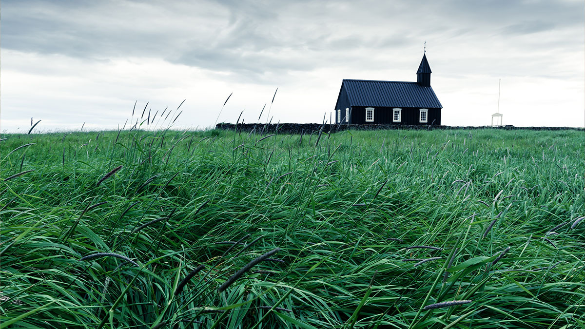 A black church building sits in the middle of an overgrown field under a stormy sky