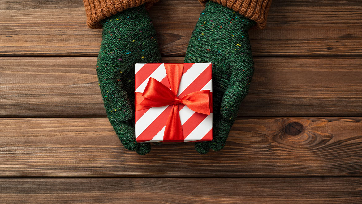 A person wearing mittens holds out a small wrapped Christmas gift