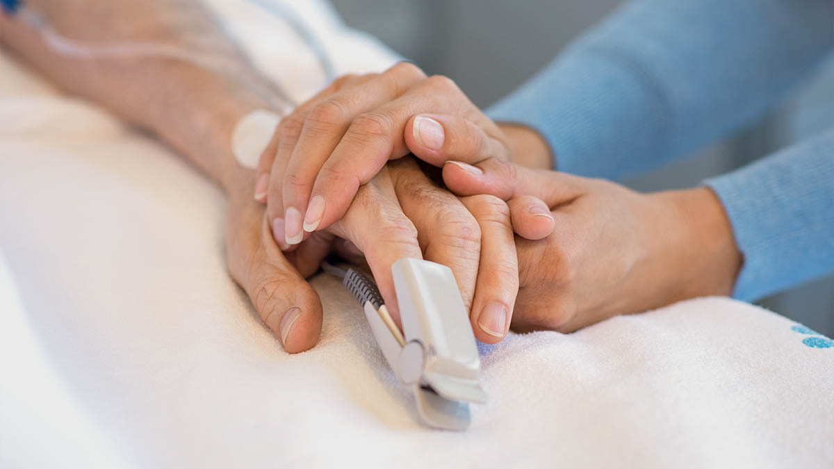 A young woman holds her elderly family member's hand in a hospital