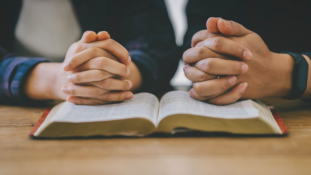 A man disciples a younger boy, and they both fold their hands on a Bible to pray together