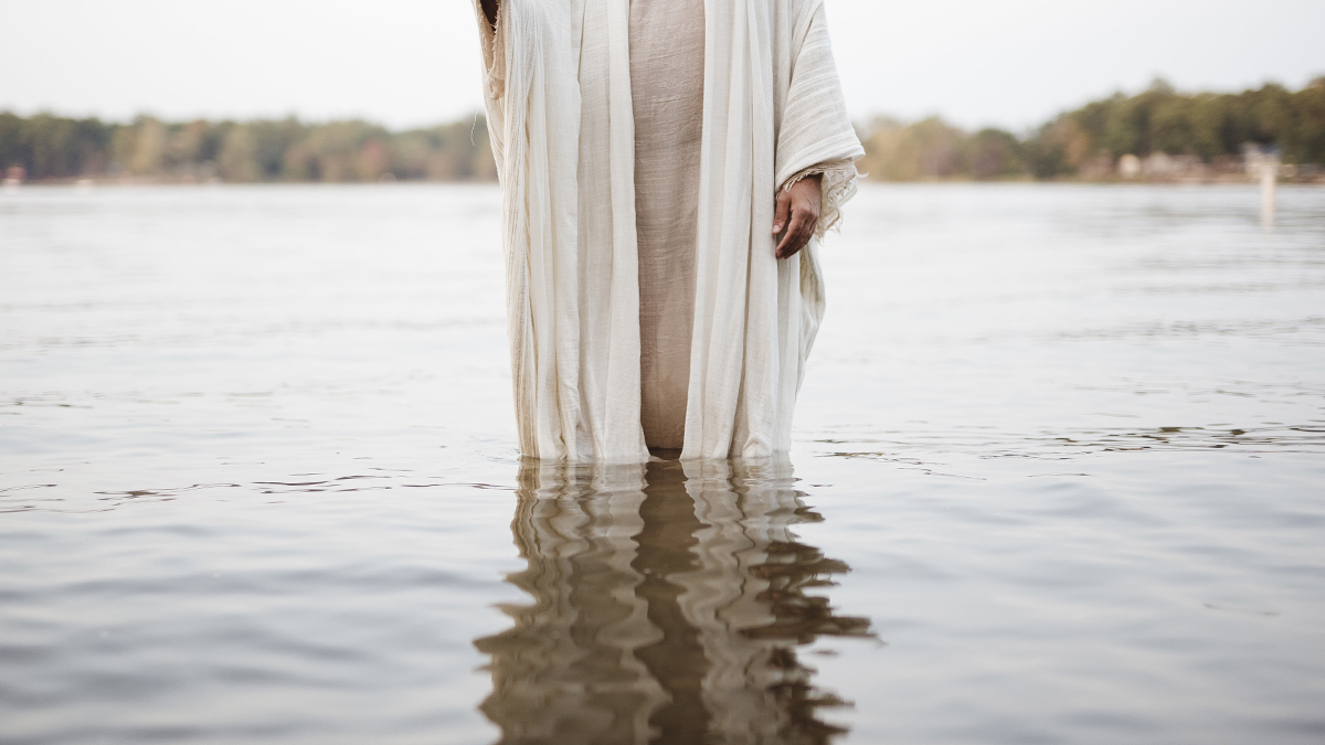 Jesus' face is hidden as he stands in a river — you can see the reflection of his robe in the water