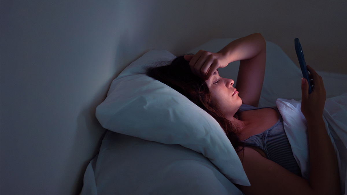 A young woman lies awake and scrolls on her phone because she is anxious and worried