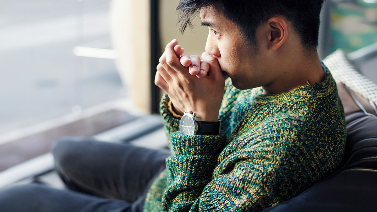 A young man folds his hands and stares out a window in thought