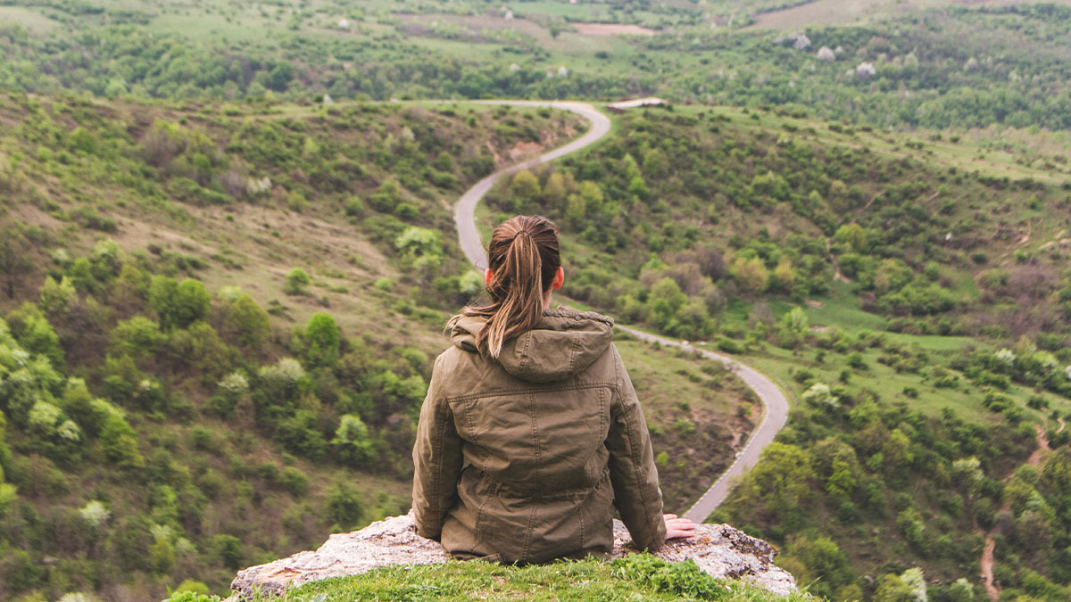 A girl sits on a cliff overlooking a road winding through a valley